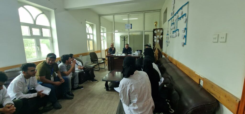 People seated in a meeting room around a central table, facing two men at the front as others listen and take notes.