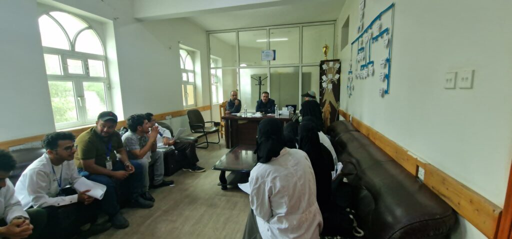 Group of people in a conference room listening to a panel at a desk; several attendees sit on chairs and couches, women in hijabs on the right.