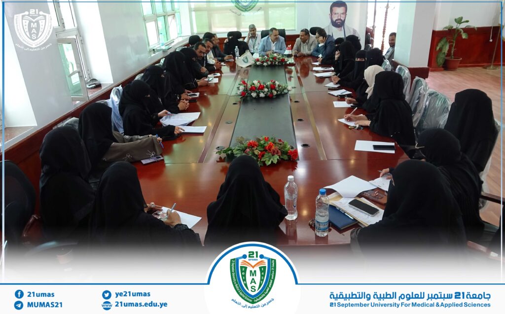 Group of people in a formal meeting around a long conference table, women in black abayas and men in shirts, with floral centerpieces in a university room.
