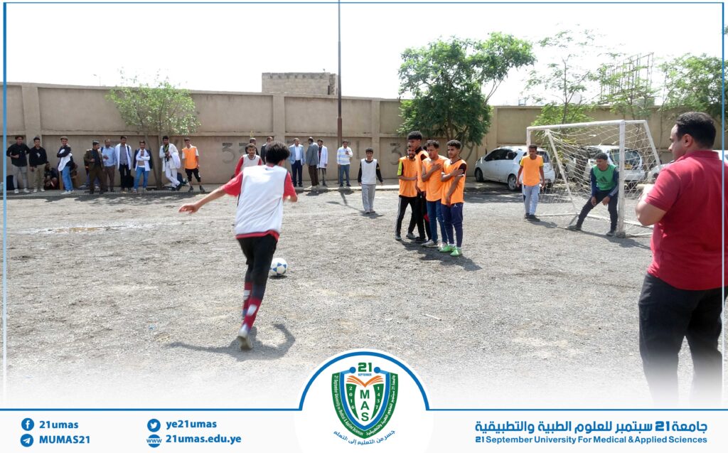 Group of young men watching a street soccer game on a dirt lot beside a tall wall; a player in white and red is kicking the ball toward the goal defended by a goalkeeper in green.