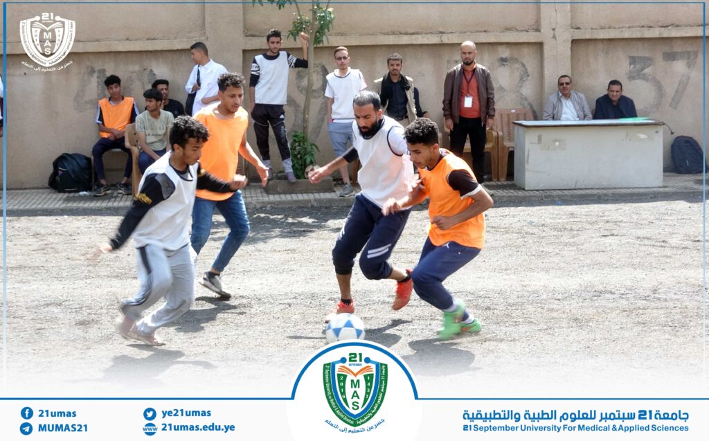 Group of young men playing outdoor soccer, wearing orange pinnies and white jerseys, with spectators along a wall watching the game; event branding for 21 September University visible at bottom.