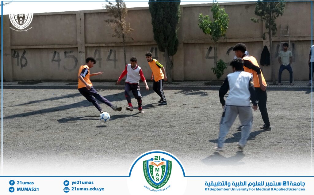Group of students playing soccer on an outdoor dusty court beside a tall wall; some players wear orange bibs and a ball is in play.