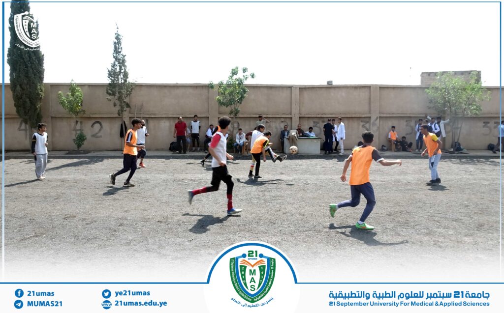 Young men playing soccer on an outdoor gravel field beside a tall beige wall, some wearing orange bibs, with spectators sitting along the wall; a university logo appears at the bottom edge.