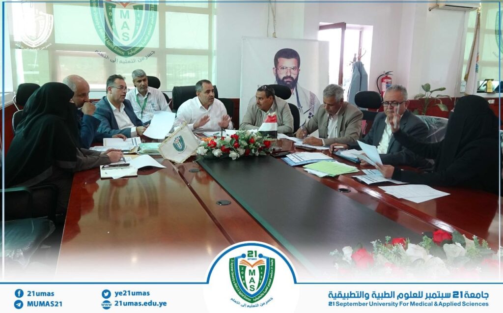 Group of officials around a conference table in a university meeting room, with the 21MAS logo and floral centerpiece visible.