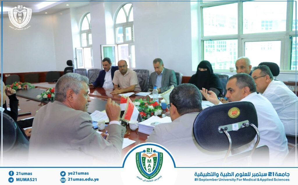 Group of professionals in a formal meeting around a long conference table in a bright room with large windows, floral centerpieces and national flags in view.