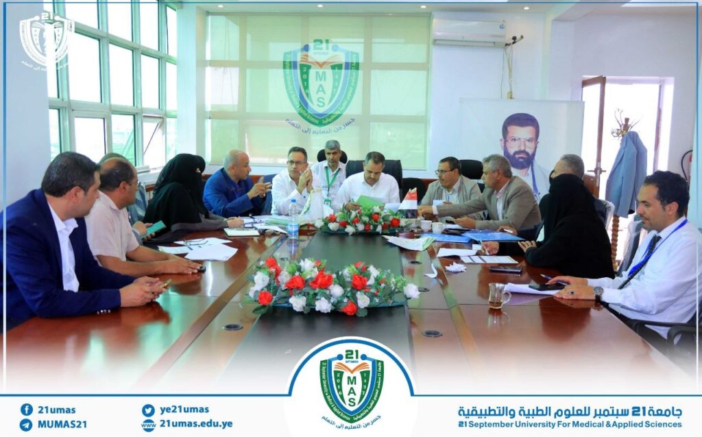 Group of professionals seated around a conference table in a meeting room of 21 September University for Medical & Applied Sciences, with a campus emblem on the glass behind them.