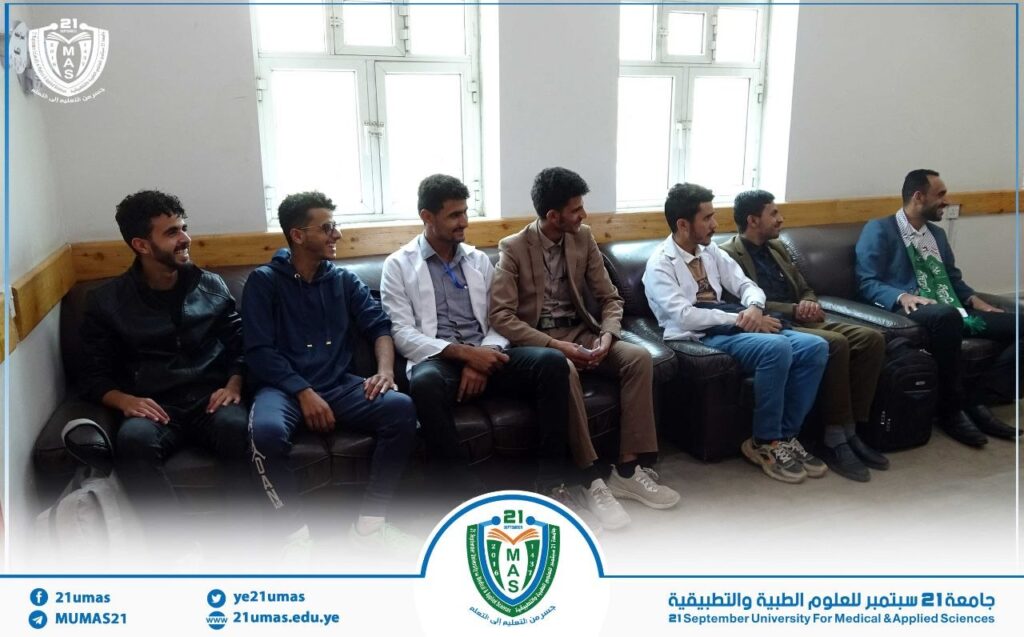 Group of seven men sitting on a brown couch in a bright room, smiling and chatting during a university gathering, with a circular logo overlay at the bottom.