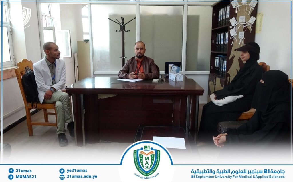 Man in a brown jacket sits at a wooden desk, flanked by a man in a light suit and two women in black hijabs, in a university office setting with a blue-bordered banner at the bottom.