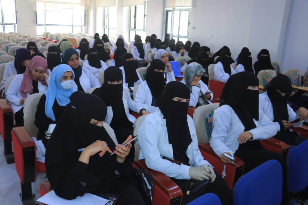 Group of women in white lab coats and hijabs seated in a classroom lecture hall, some looking at phones or notes.