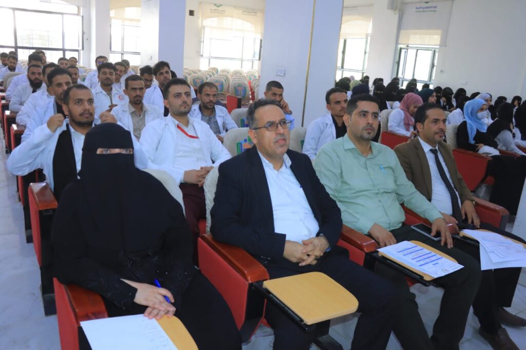 Audience seated in a conference hall listening to a presentation; woman in black niqab in the foreground.