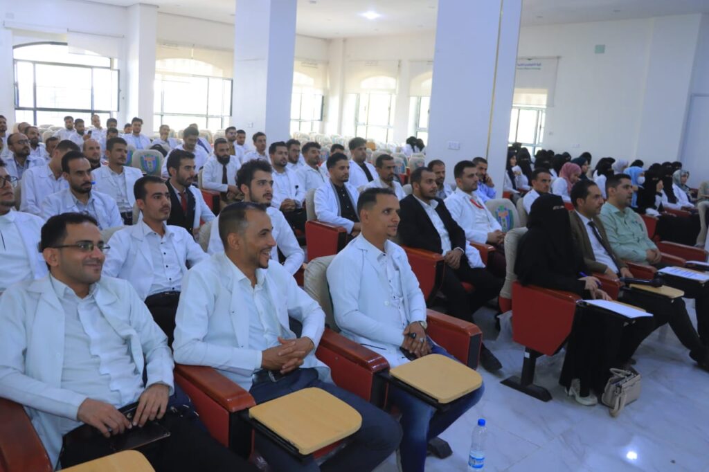 Audience of doctors and students seated in a bright lecture hall, many in white coats and some in hijabs, listening to a presentation.