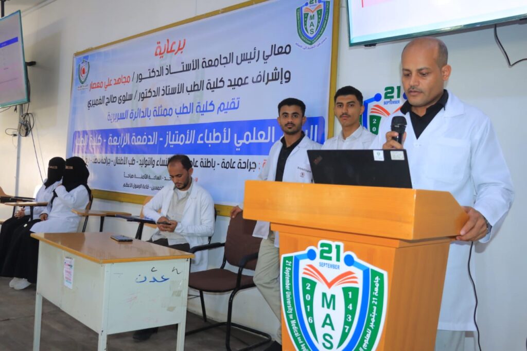 Man in a white coat speaks at a wooden podium with a MAS badge, as others in white coats sit behind and a large banner with Arabic text hangs in the background.