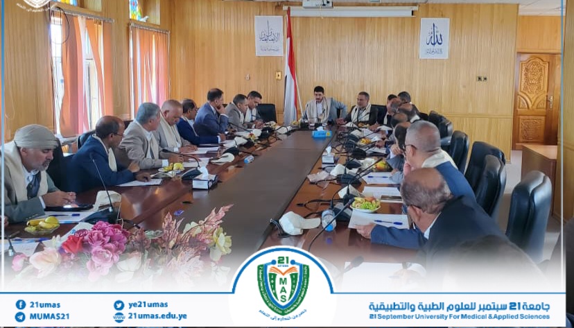 Formal board meeting with about 20 people seated around a long conference table in a wood-paneled room, with flags and banners in the background.
