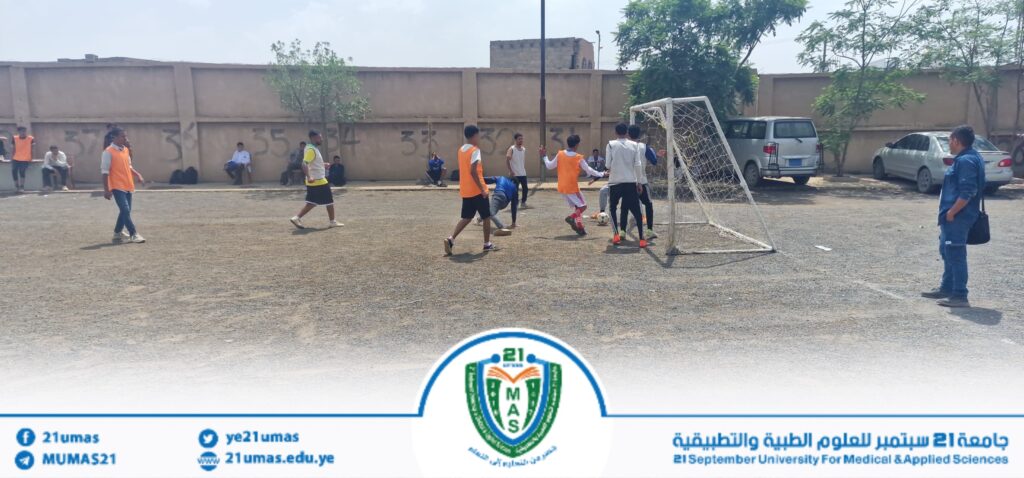 Group of young men playing soccer on a gravel field near a wall, some wearing orange vests, with a small goal and spectators seated along the wall in the background.