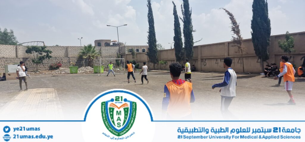 Group of students playing soccer on a dusty outdoor court beside a tall wall, some in orange vests and others watching.