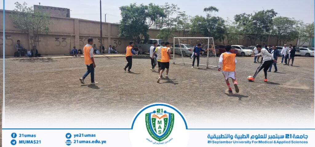 Group of students playing soccer on a dusty field, orange bibs, a goalie near a small goal, and spectators along a wall with numbers in the background, campus setting.
