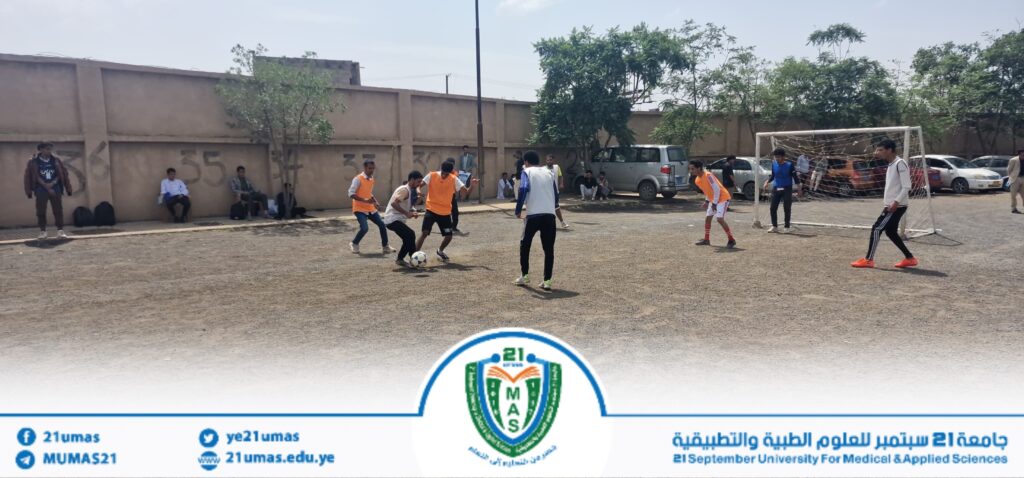 Group of men playing soccer on a dusty outdoor field beside a tall wall, with players in orange bibs chasing the ball near a goal and spectators along the wall.