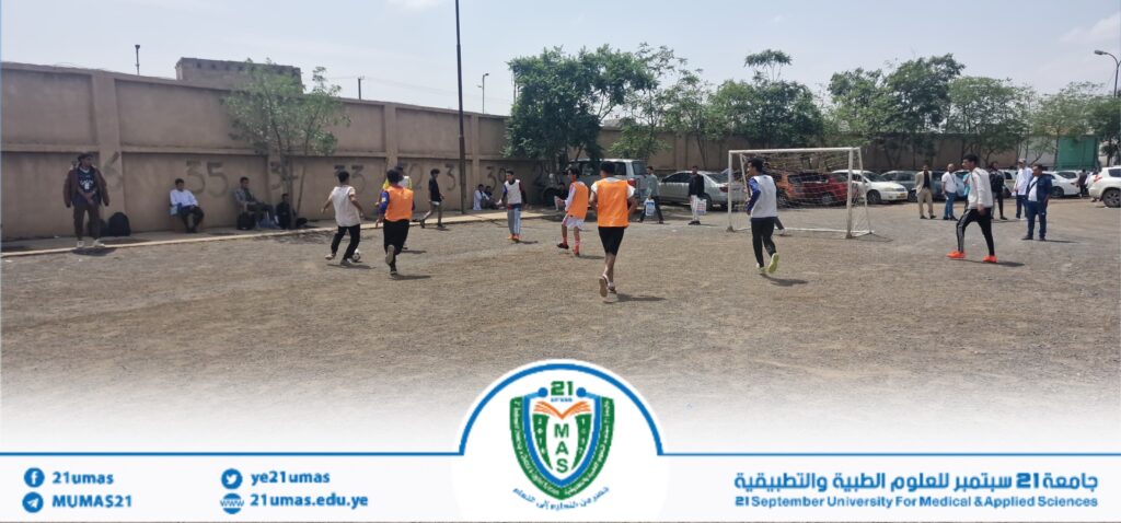 Group of young men playing a soccer match on a dusty outdoor field, orange bibs, goalpost on the right, with a wall and parked cars in the background.