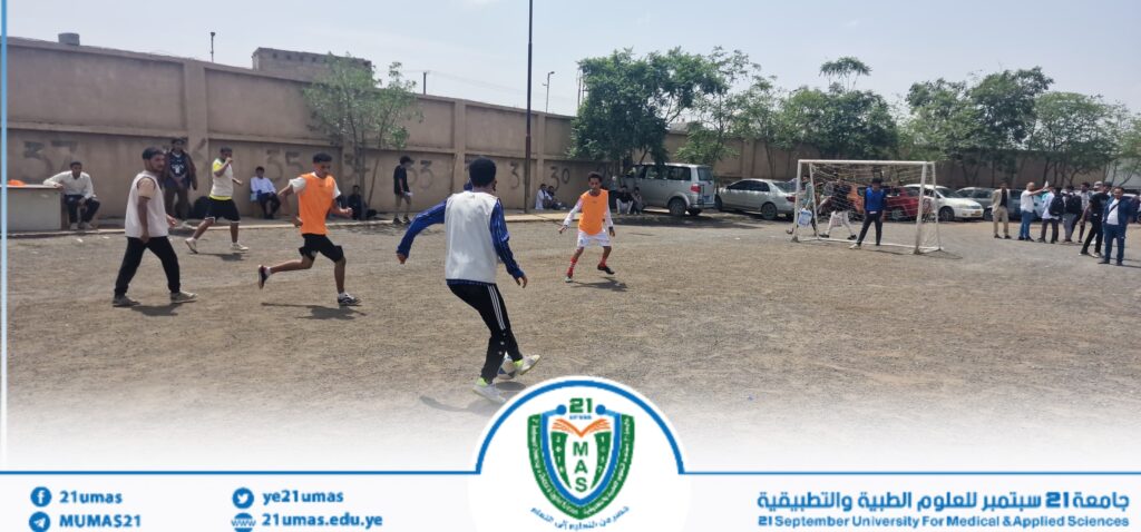 Young men playing a street soccer game on a dirt field near a low wall, with a small goal and onlookers along the sidelines; banner at the bottom shows a university logo.