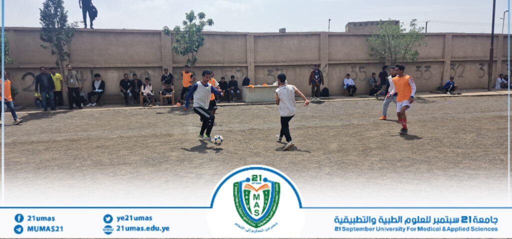 Youth soccer match on a dusty field with players in orange pinnies; spectators sit and stand along a concrete wall, while a university banner is visible at the bottom.