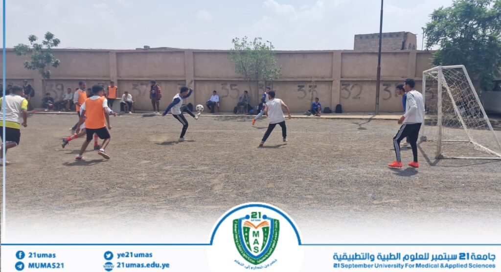Young men playing soccer on a dirt field, a ball mid-kick near the goal, with spectators along a wall in the background and a campus logo at the bottom edge