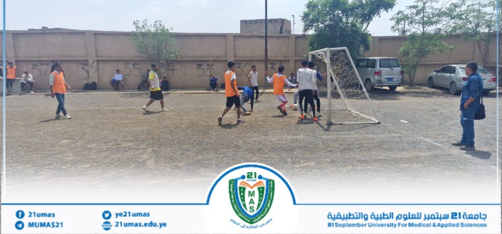 Group of students playing soccer on a dusty field near a wall, with some players in orange vests and a small goal.