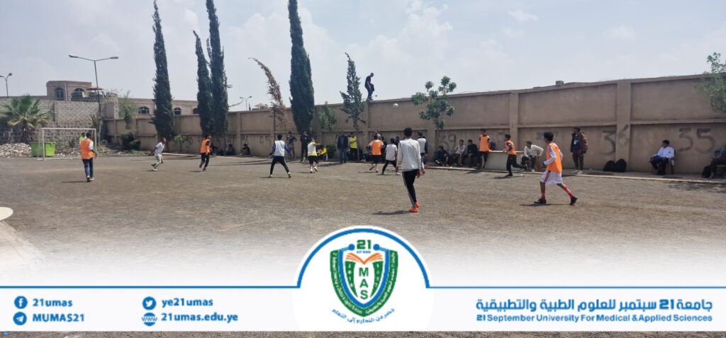 Groups of young players on a dusty outdoor court, some in orange vests, kicking a soccer ball with a wall and trees behind them.