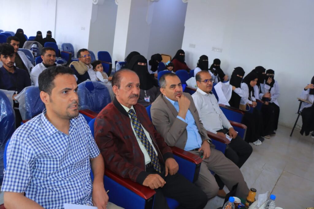 Audience in a conference room; front row has three men in shirts/jackets, back rows include women in black hijabs.