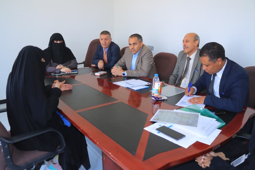 1001235745 - جامعة 21 سبتمبر Five men in suits and two veiled women sit around a conference table with documents and a water bottle on the table in a formal meeting.