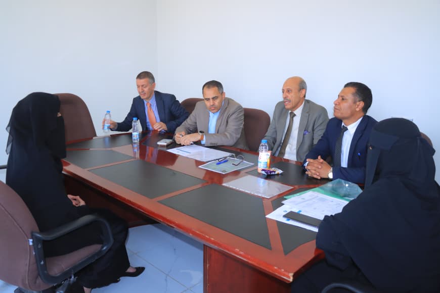 1001235746 - جامعة 21 سبتمبر Five men in suits and two women in black abayas sit around a rectangular conference table during a business meeting.