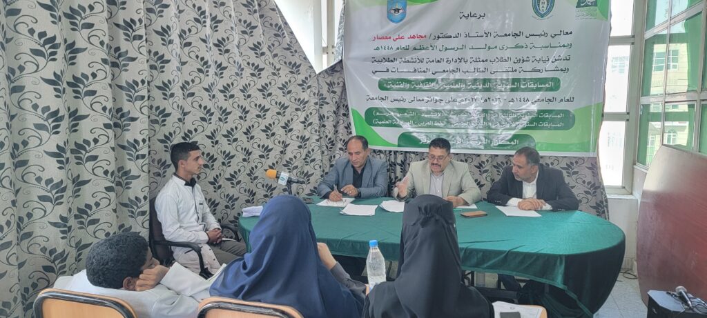 Three men sit at a green-covered panel table speaking into a microphone, with an Arabic banner behind them.