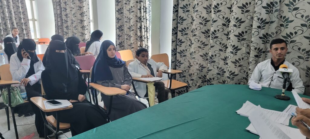 Group of students in a classroom listening to a speaker at a table with a microphone on the right.
