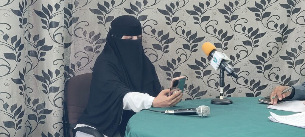 Woman in a black niqab seated at a table, holding a smartphone, with a yellow-muffed microphone in front for a press recording.