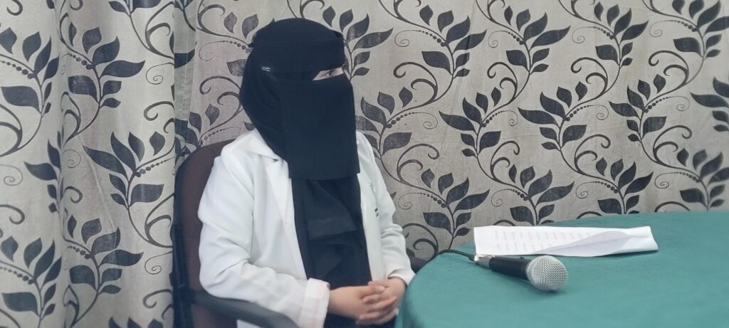 Person wearing a black niqab and white lab coat sits at a table with papers and a microphone during a talk