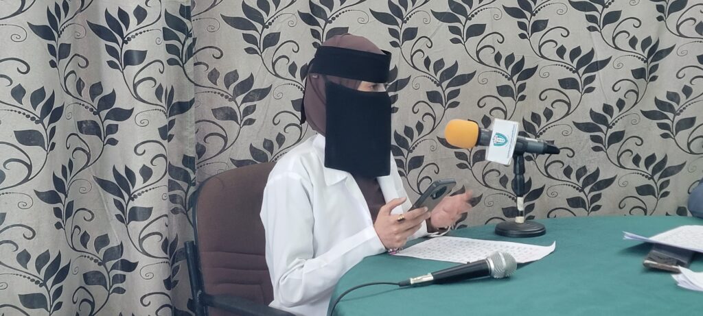 Woman wearing a brown veil and black niqab seated at a table with a microphone, papers, and a phone during a press briefing/meeting.