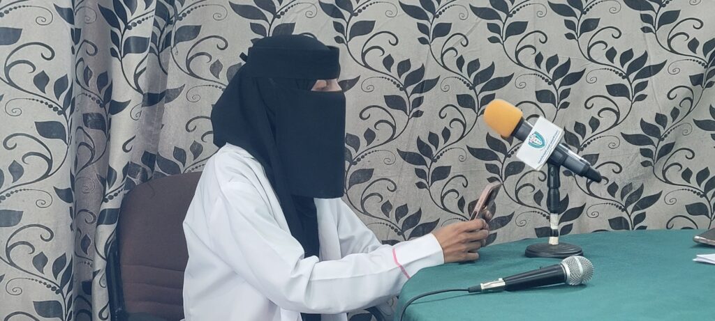 Person wearing a black niqab and white coat sits at a table with microphones, giving a press-style interview.