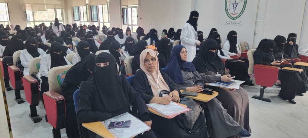 Large lecture hall filled with women wearing hijabs and niqabs, some in white coats, taking notes or listening attentively.