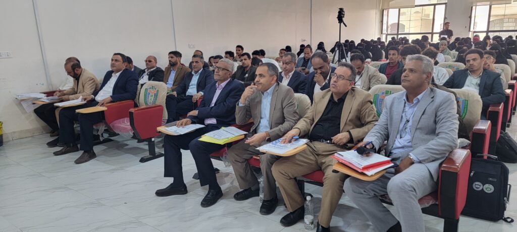 Audience seated in a conference hall, front row in suits taking notes while a camera stands on a tripod in the back.