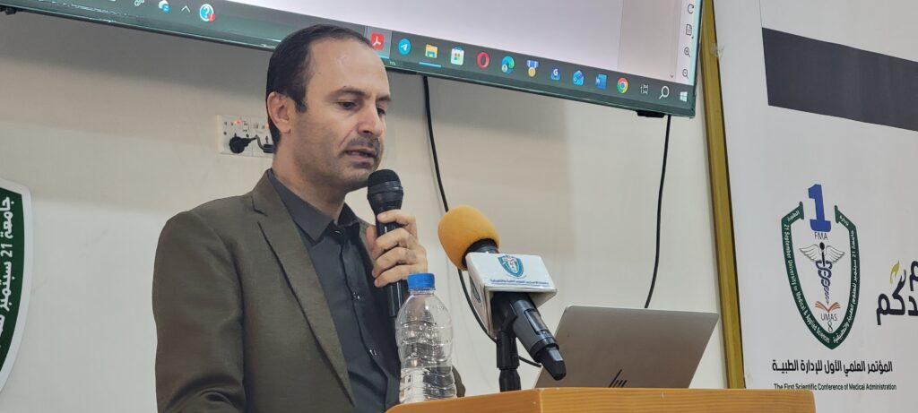 Man in a brown suit speaks into a handheld microphone at a conference podium, with a laptop and water bottle nearby, and a banner with Arabic text and logos in the background.