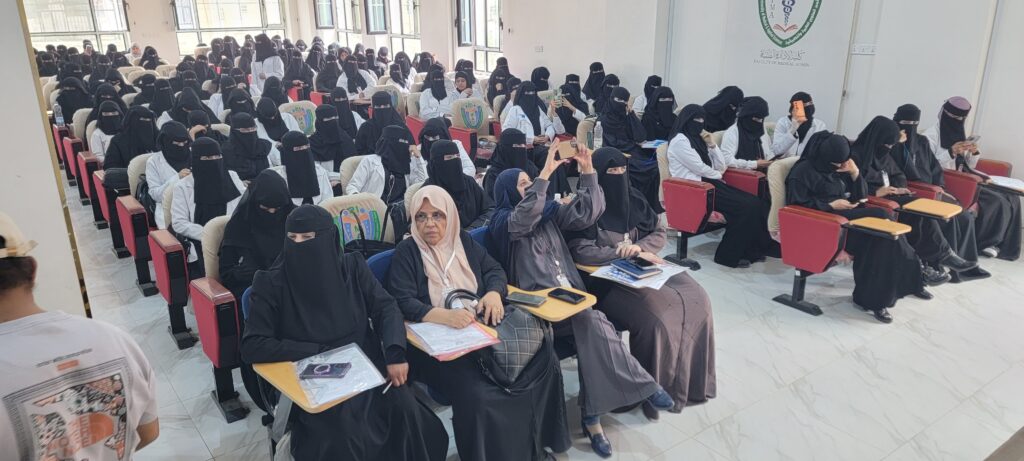 Auditorium filled with female medical students wearing niqab/covering attire, seated with notebooks and phones for a lecture.