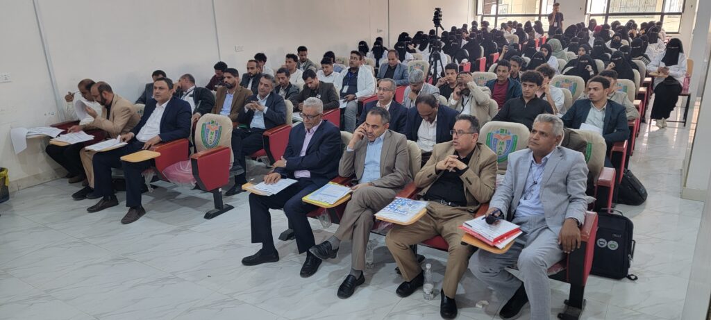 Audience attending a formal seminar in a large lecture hall; front row in suits, back rows with students wearing hijabs and masks.