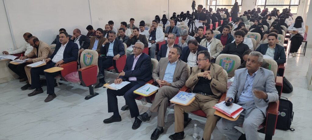 Audience in a lecture hall: front row of men in suits taking notes, rows of attendees behind them listening to a presentation.