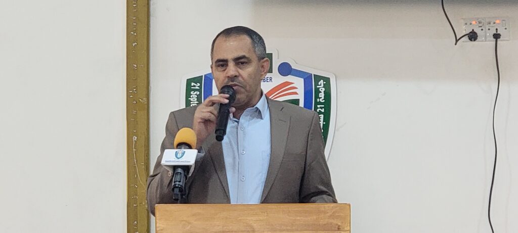 Man in a brown suit speaks into a handheld microphone at a wooden podium, delivering a talk indoors with a crest behind him.