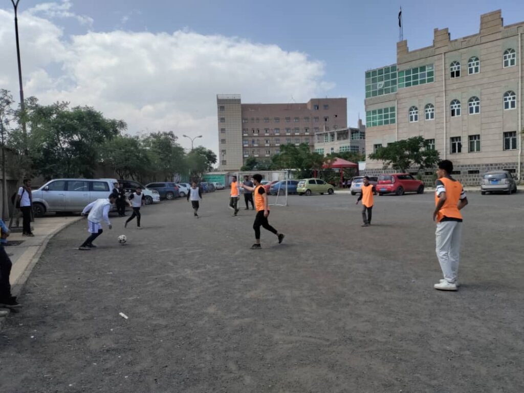 Group of youths playing soccer on a gravel lot beside parked cars, some wearing orange training vests, with buildings in the background.