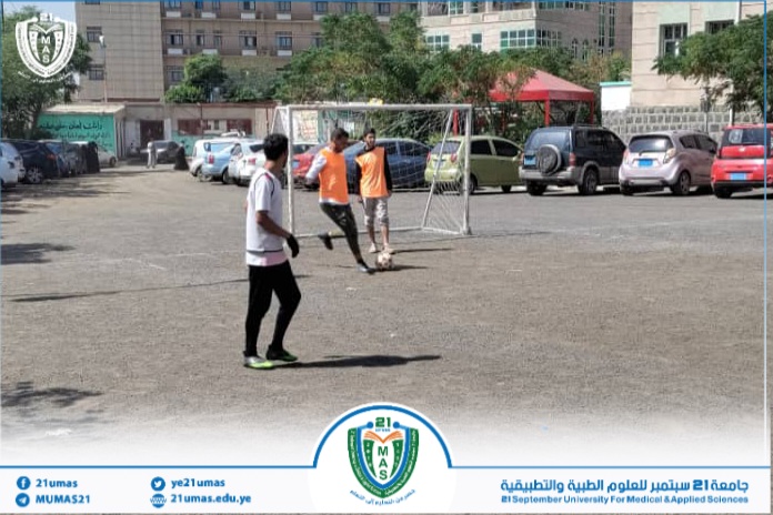 Students playing soccer on an outdoor campus field with a goal; parked cars and buildings in the background, university logo at the bottom.