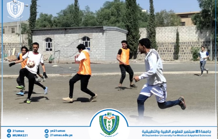 Group of men playing soccer on an outdoor dusty court; one player in white kicks the ball while others in orange vests run after it, with buildings, a stone wall and trees in the background and a university crest/banner at the bottom.