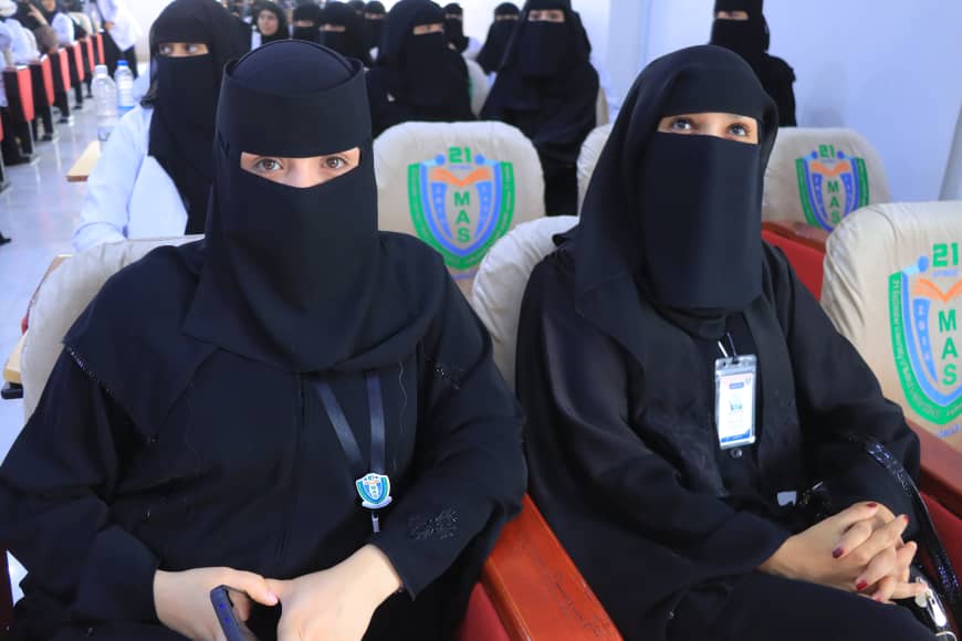 Two women in black abayas and niqabs sit in a conference hall wearing badges; audience members are visible behind them.