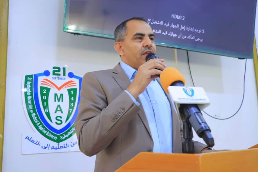 Man in a tan suit speaks into an orange microphone at a podium; a school crest with 21 September MAS is on the wall behind him.