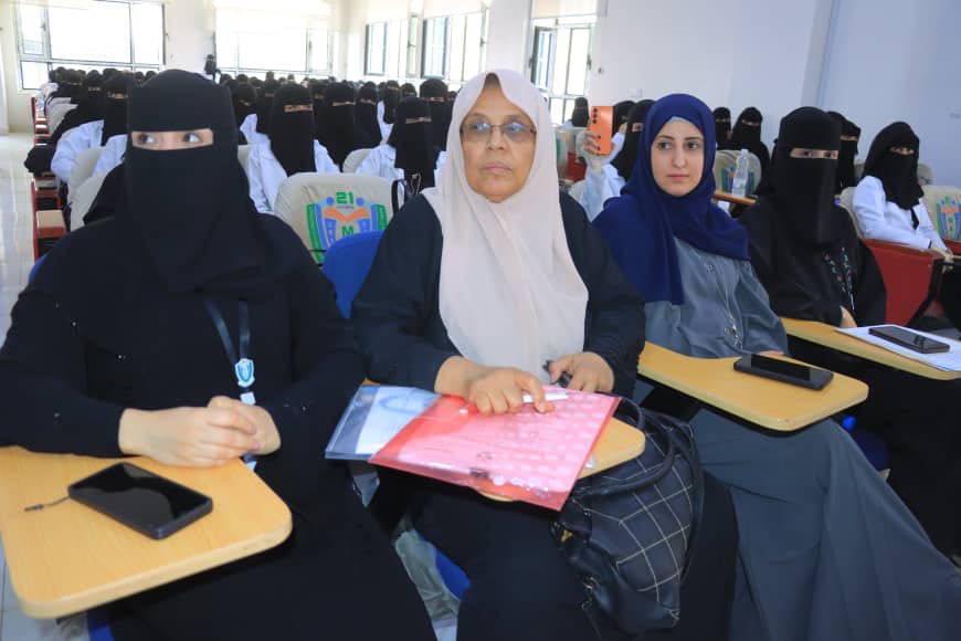 Front-row women in a classroom: left in a black niqab, middle in a beige headscarf holding a pink folder, right in a blue hijab; rows of students in hijabs in the background.