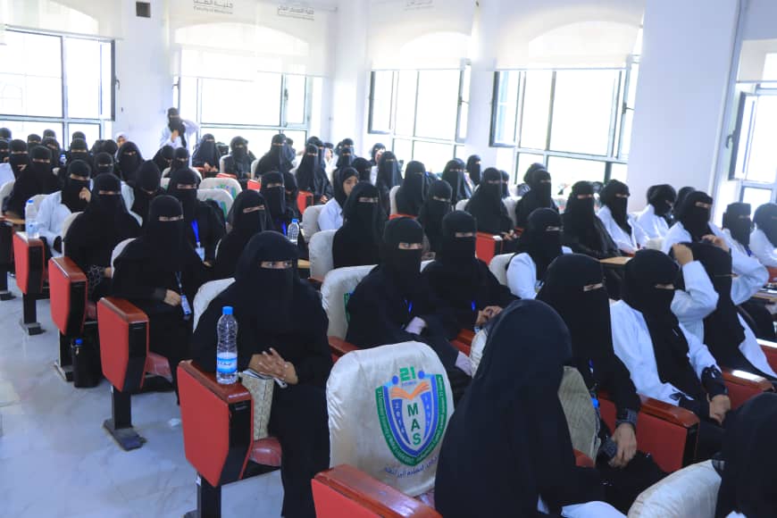 Large lecture hall filled with women wearing black niqabs and hijabs seated in red chairs, listening to a lecture with water bottles on desks nearby.